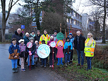 Gruppenbild an der Straße vor der Schule mit einer der Holzfiguren, einem Mädchen, das ein Schild mit der Aufschrift 