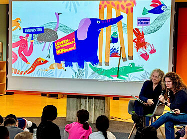 Auf der Leinwand ist ein Bild mit Tieren aus dem Dschungel zu sehen, davor sitzen die beiden Vorleserinnen.