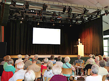 Vorne das Publikum, auf der Bühne Thomas Hofschuster hinter einem Rednerpult Vorne das Publikum, auf der Bühne Thomas Hofschuster hinter einem Rednerpult