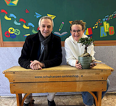  Norbert Seidl und Heike Senzel sitzen auf einer Schulbank, Heike Senzel hält das Bonsaibäumchen.